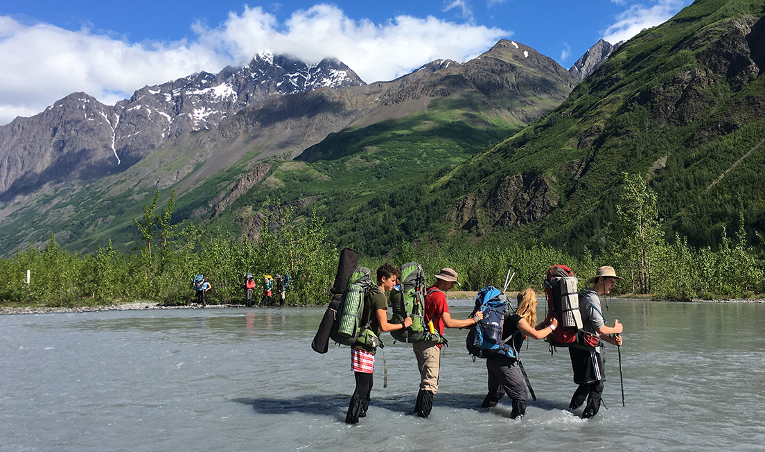 teenagers crossing a river in alaska with backpacks