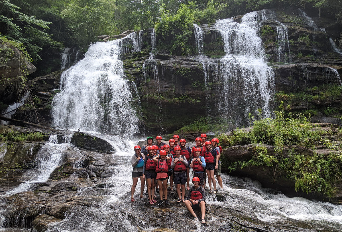 teenage boys and girls wearing life jackets standing in front of a waterfall